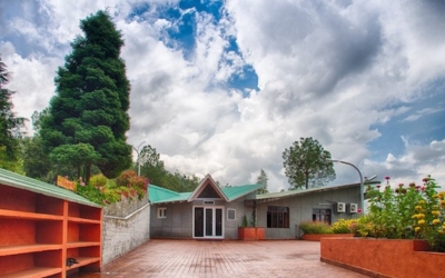 Modern, small ashram building with a large courtyard and surrounding flower garden under a cloudy sky